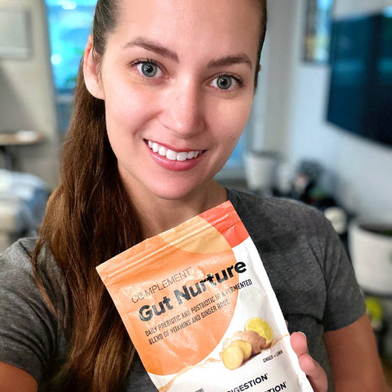 A person holding a packet labeled Complement Gut Nurture, which features images of ginger and lemon slices. They are smiling and wearing a gray shirt. The background is a blurred indoor setting.