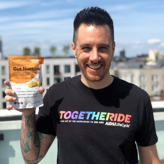 A person with short hair and tattoos smiles while holding a packet labeled Gut Nurture on a rooftop. Theyre wearing a black T-shirt with the words TOGETHER RIDE in colorful letters. The sky and buildings are visible in the background.