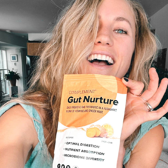 A person smiling and holding a package of Complement Gut Nurture, which is a prebiotic and postbiotic blend. The package highlights benefits like optimal digestion, nutrient absorption, and microbiome diversity. The person is indoors.