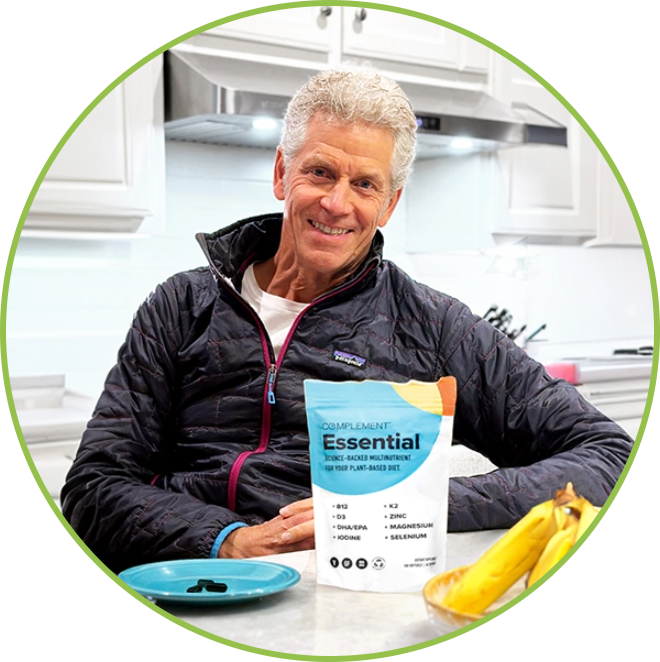 A person with gray hair sits in a kitchen, smiling. They are wearing a black jacket and are next to a package labeled Complement Essential, which is on the counter beside a small teal plate and some bananas.