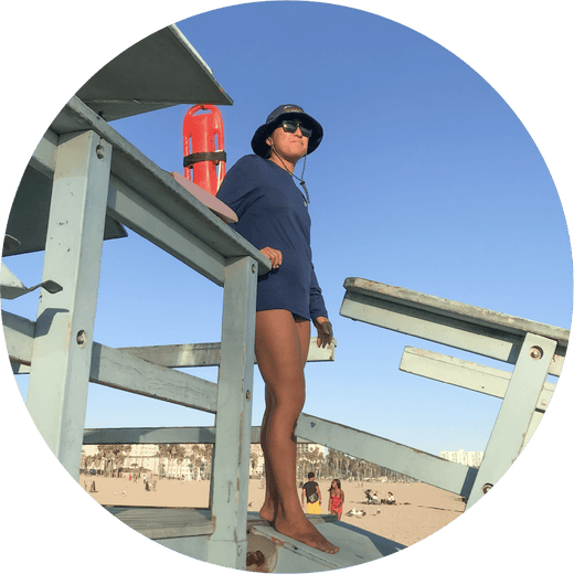 A lifeguard stands on a wooden lifeguard tower, wearing a blue uniform and hat, with a rescue float nearby. The beach and a few beachgoers are visible in the background under a clear blue sky.