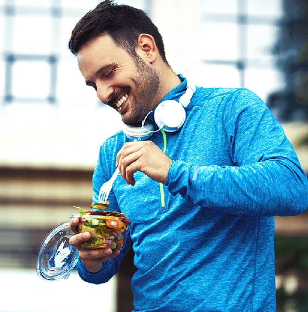 A man in a blue long-sleeve shirt smiles while eating a salad. He has white headphones around his neck and holds a fork in one hand and the container in the other. The background is slightly blurred.