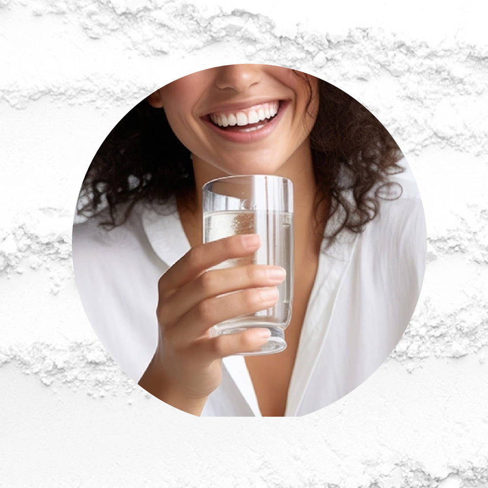 A woman with curly hair and a bright smile holds a glass of water up to her mouth. She wears a white shirt, and the background features a textured white surface.