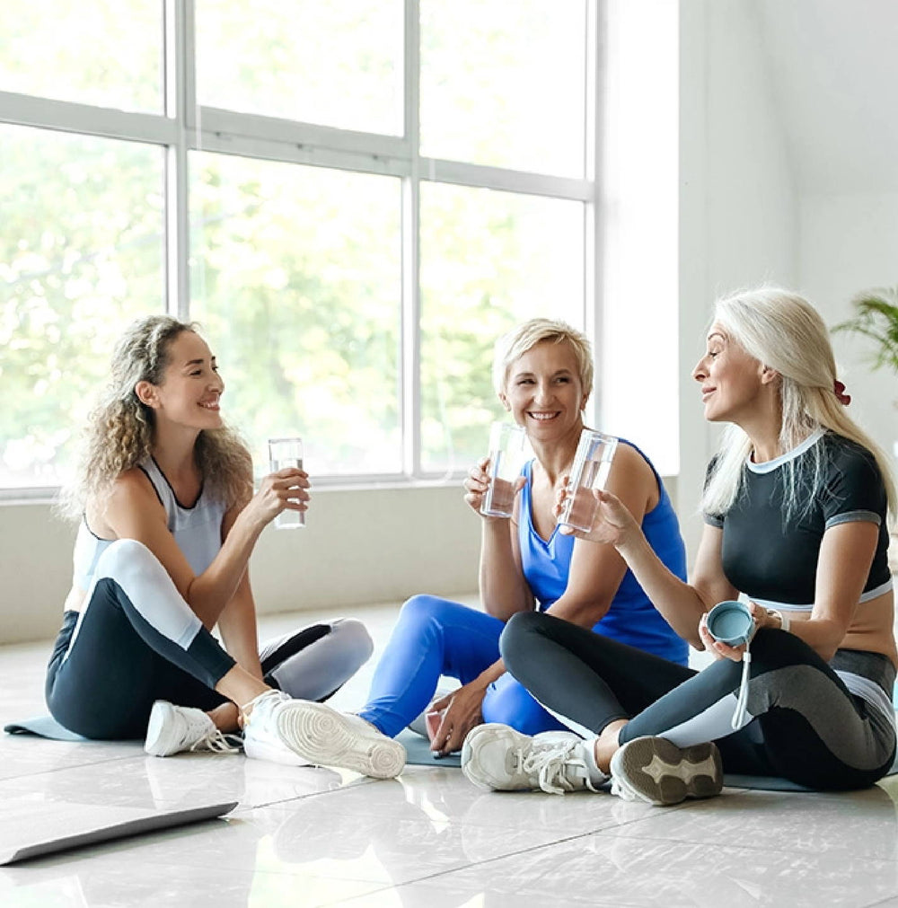 Three women sit on a gym floor, smiling and holding water bottles. They wear athletic attire and seem relaxed after exercising. Large windows in the background let in natural light, creating a bright and cheerful atmosphere.