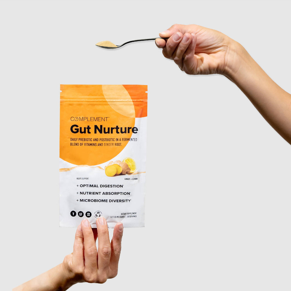 A close-up of a hand holding a Complement Gut Nurture supplement bag, featuring a fermented blend of vitamins and ginger root. Another hand holds a spoon with powder above the bag. The background is plain grey.