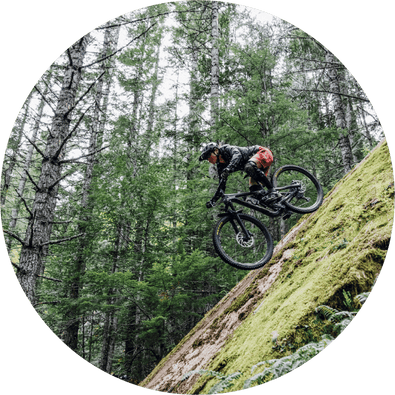 A mountain biker wearing a helmet and red shorts rides down a steep, moss-covered slope in a dense forest. Tall trees surround the area, creating a lush, green backdrop.