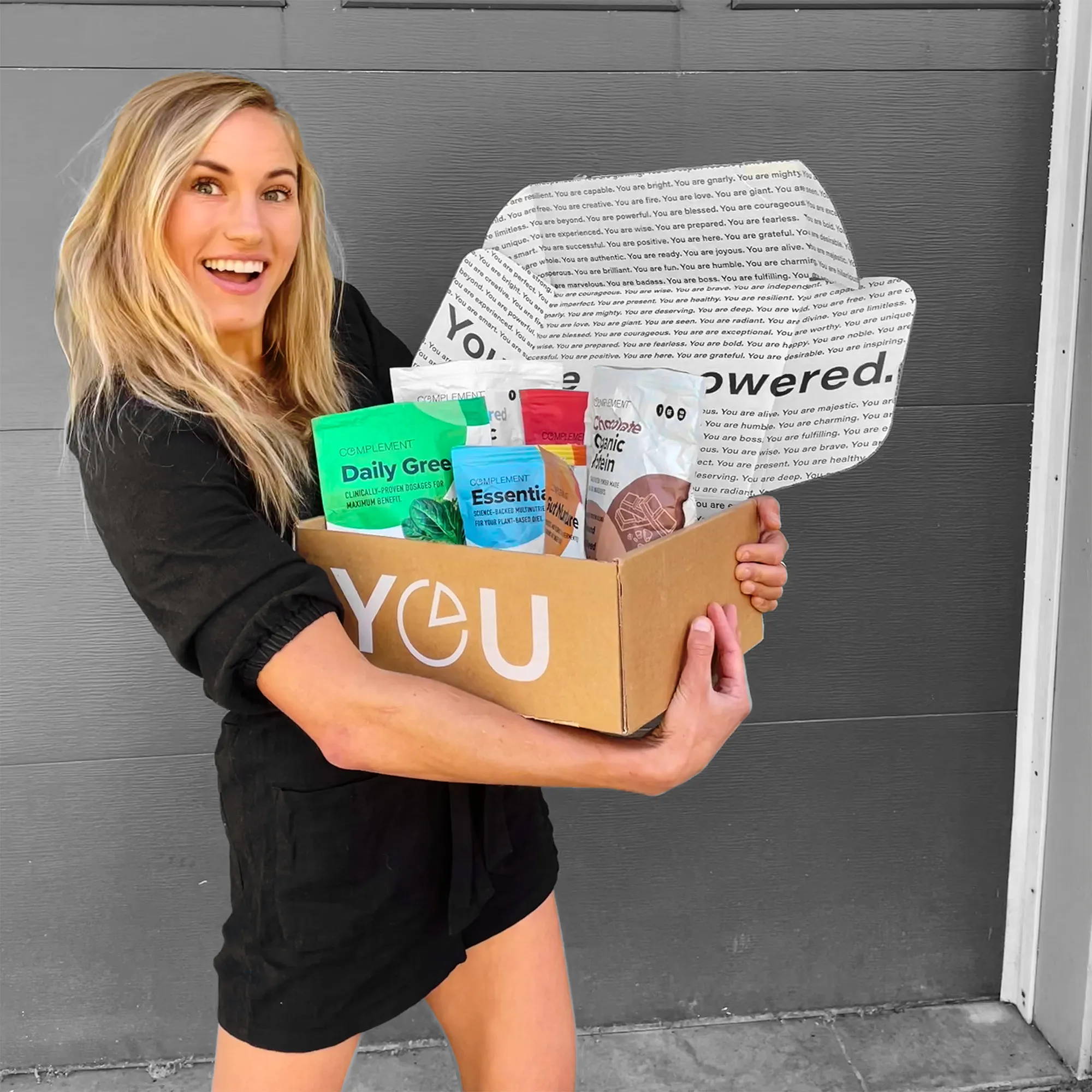 A smiling woman stands in front of a gray door, holding an open YEU-branded box filled with various health products, including Daily Greens and Essential Protein packets. She is wearing a black outfit and has long blonde hair.