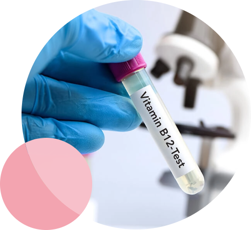 A gloved hand holds a test tube labeled Vitamin B12 Test. The tube has a pink cap and contains a light-colored liquid. Blurred laboratory equipment is visible in the background.