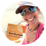 A person smiling outdoors, wearing a pink tank top, sunglasses, and a colorful cap, holds a packet labeled Gut Nurture with details about prebiotics and postbiotics. The background features a natural setting with water.