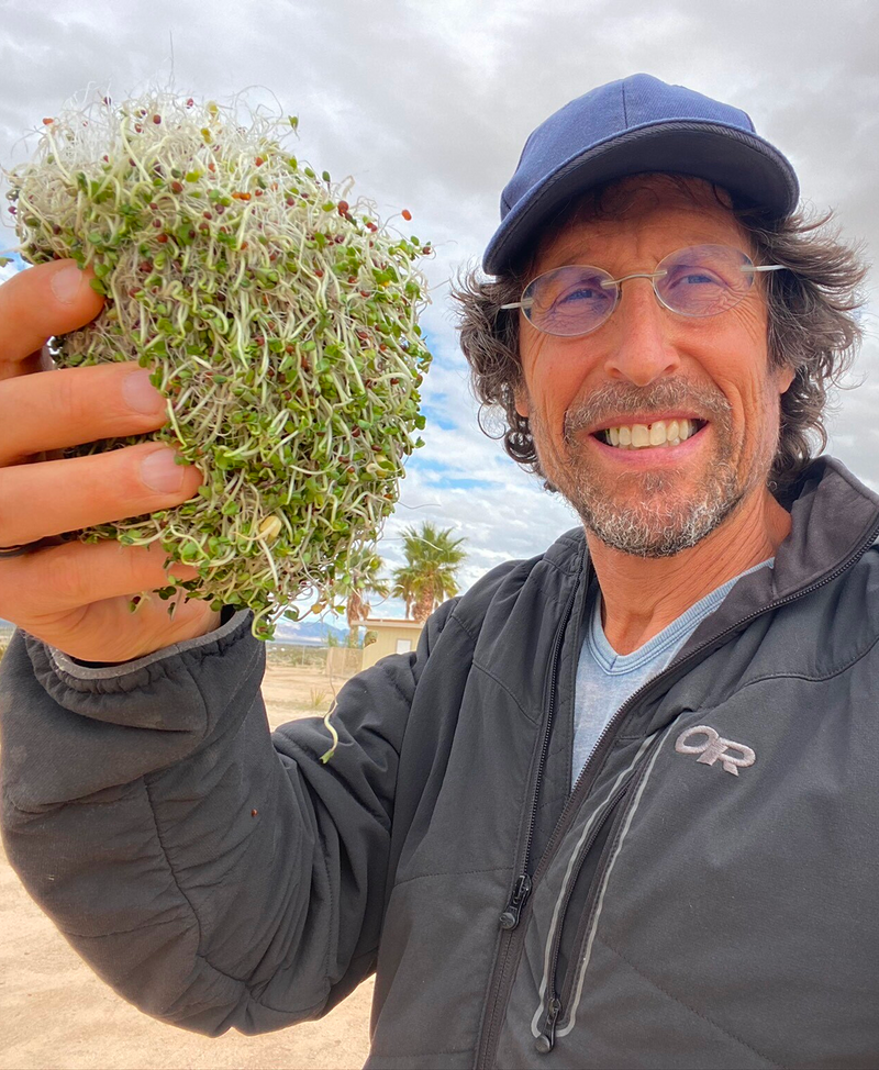 A man wearing glasses, a blue cap, and a black jacket is smiling and holding a large handful of fresh sprouts outdoors with a cloudy sky and palm tree in the background.