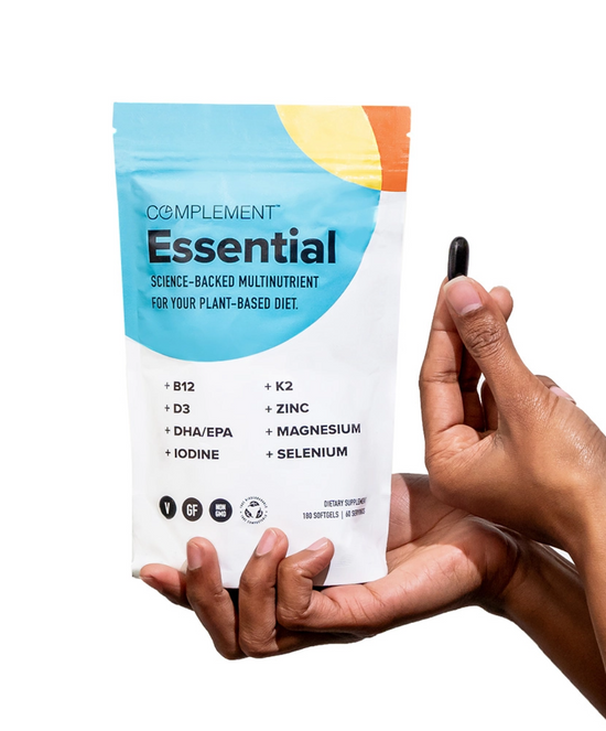 A person holding a black capsule next to a Complement Essential multinutrient pouch, which lists nutrients like B12, D3, DHA/EPA, iodine, K2, zinc, magnesium, and selenium. The packaging is white with blue and orange accents.