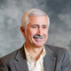 A middle-aged man with short gray hair and a mustache, wearing a gray blazer and a light-colored shirt, smiling against a blurred gray background.