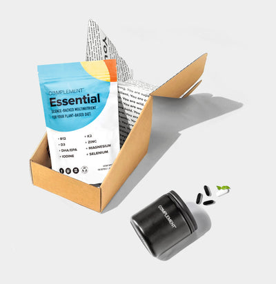 A cardboard box is open, displaying a packet labeled Essential in blue and yellow. Next to it is a black container labeled COMPLEMENT, with several black pills and a green capsule beside it.