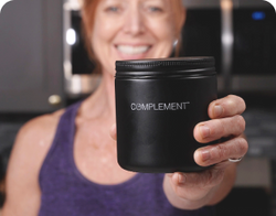 A person in a purple tank top holds a black jar labeled COMPLEMENT towards the camera, smiling. The background is blurred, focusing on the jar.