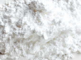 A close-up view of a pile of fine white powder with a fluffy, soft texture, resembling baking ingredients such as powdered sugar or cornstarch, spread on a white surface.