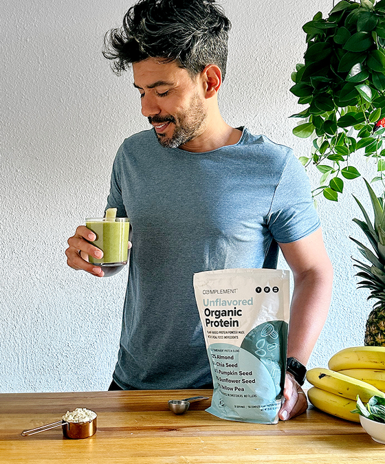 A man in a light blue shirt holds a green smoothie and stands by a wooden table with a bag of Complement Organic Protein powder, measuring scoop, bananas, and a leafy plant in the background.