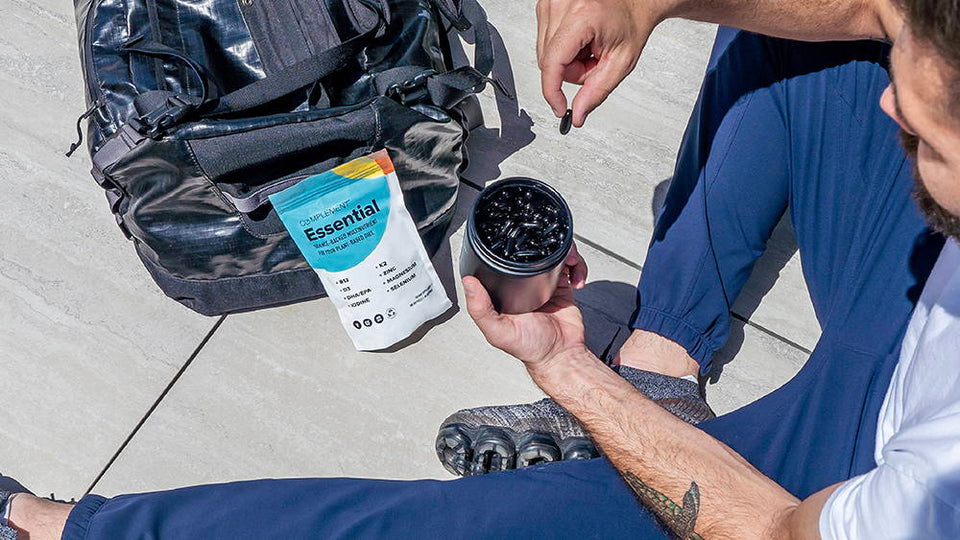 A person sitting on a tiled floor holds a container with black capsules. A bag is nearby with a pouch labeled Essential displaying nutrition facts. The person wears blue pants, a white shirt, and gray shoes.