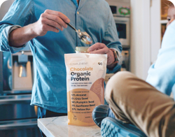 A person scoops powder from a bag of Complement Chocolate Organic Protein on a kitchen counter. The bag lists ingredients like almond, chia seed, pumpkin seed, and more. Another person sits nearby with feet up.