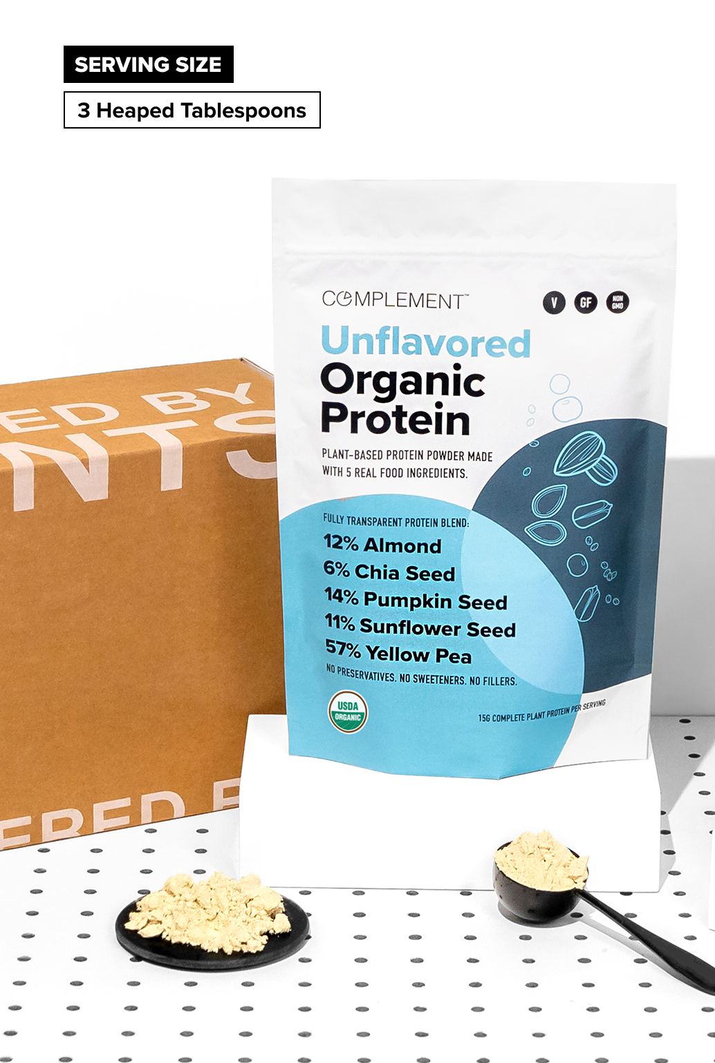 A bag of Complement Unflavored Organic Protein powder stands upright. In front, a black spoon with protein powder sits on a white block. A brown box and dotted surface are in the background. Text reads: 3 Heaped Tablespoons Serving Size.