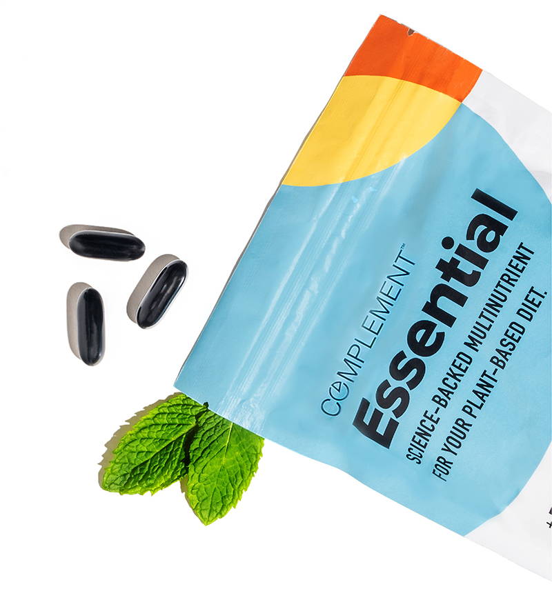 A blue and orange resealable pouch labeled Complement Essential with text highlighting plant-based nutrition. Four capsules lie beside the bag, next to sprigs of fresh mint leaves.