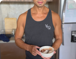 A person in a sleeveless black shirt is holding a bowl of oatmeal topped with various seeds and nuts, standing in a kitchen. The background includes a wooden cabinet and a fridge.