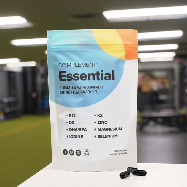 A white pouch labeled Complement Essential sits on a table in a gym. The package highlights plant-based multinutrients and lists ingredients like B12, D3, K2, zinc, and magnesium. Three black capsules are nearby.