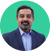 A man with a beard and mustache, wearing a dark blue suit jacket and light blue shirt, stands against a solid green background. He has short, dark hair and a neutral expression.