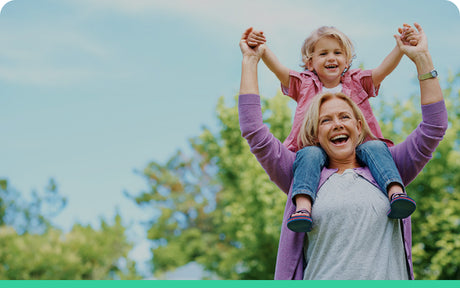 A joyful elderly woman with short blonde hair carries a young child on her shoulders. They are outdoors, surrounded by green trees and a clear blue sky. Both are smiling broadly, and the child is holding the womans hands.