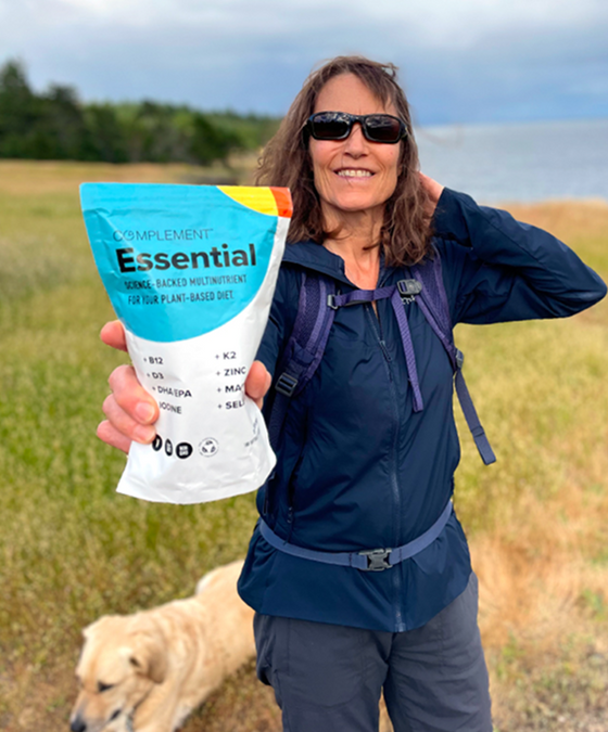 A smiling woman in sunglasses and a blue jacket holds a bag labeled Essential multivitamin outdoors, with grassy fields, water, and a dog in the background.