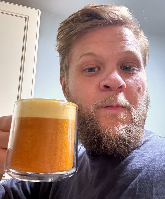 A bearded person with light brown hair holds up a glass mug filled with a frothy, orange-brown beverage, smiling slightly while standing indoors.