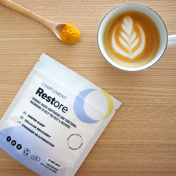 A cup of latte with a leaf-shaped design sits on a wooden table beside a packet of Complement Restore supplement, a wooden spoon, and a small pile of yellow powder.