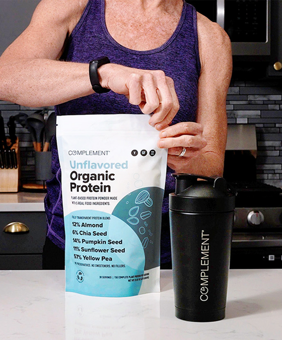 A person in a purple tank top opens a bag of Complement Unflavored Organic Protein powder in a kitchen, with a black Complement shaker cup on the counter nearby.