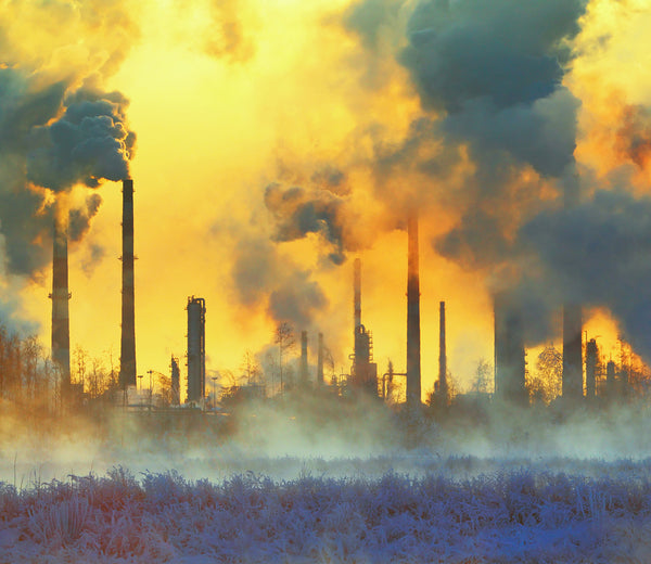 Factory smokestacks emit thick smoke into a yellow-orange sky at sunset, with dense steam and frost-covered ground in the foreground, highlighting industrial air pollution.