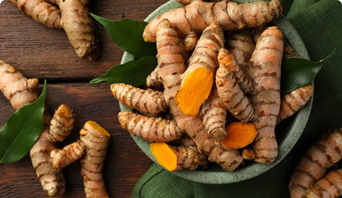 A bowl filled with fresh turmeric roots, some sliced to reveal their bright orange interior, sits on a wooden table with green leaves and a green cloth nearby.