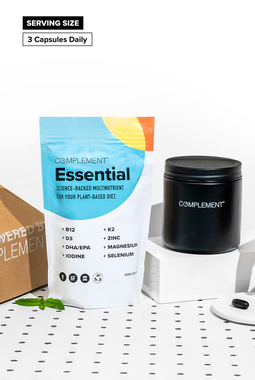 A pouch labeled “Complement Essential” and a black container sit on a white surface with a dotted pattern. The pouch lists nutrients like B12, D3, K2, DHA/EPA, iodine, zinc, magnesium, and selenium.