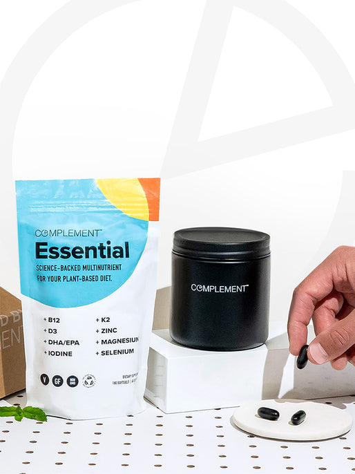 A hand picks up a black capsule near a black jar and colorful pouch of Complement's COMPLEMENT® Essential, formulated for plant-based diets with nutrients like B12, D3, omega-3 DHA/EPA, zinc, and magnesium.
