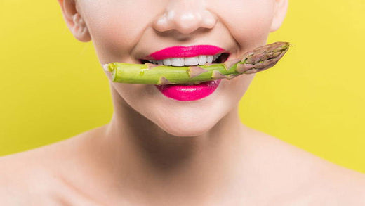 A woman with bright lipstick holds a piece of asparagus in her teeth.