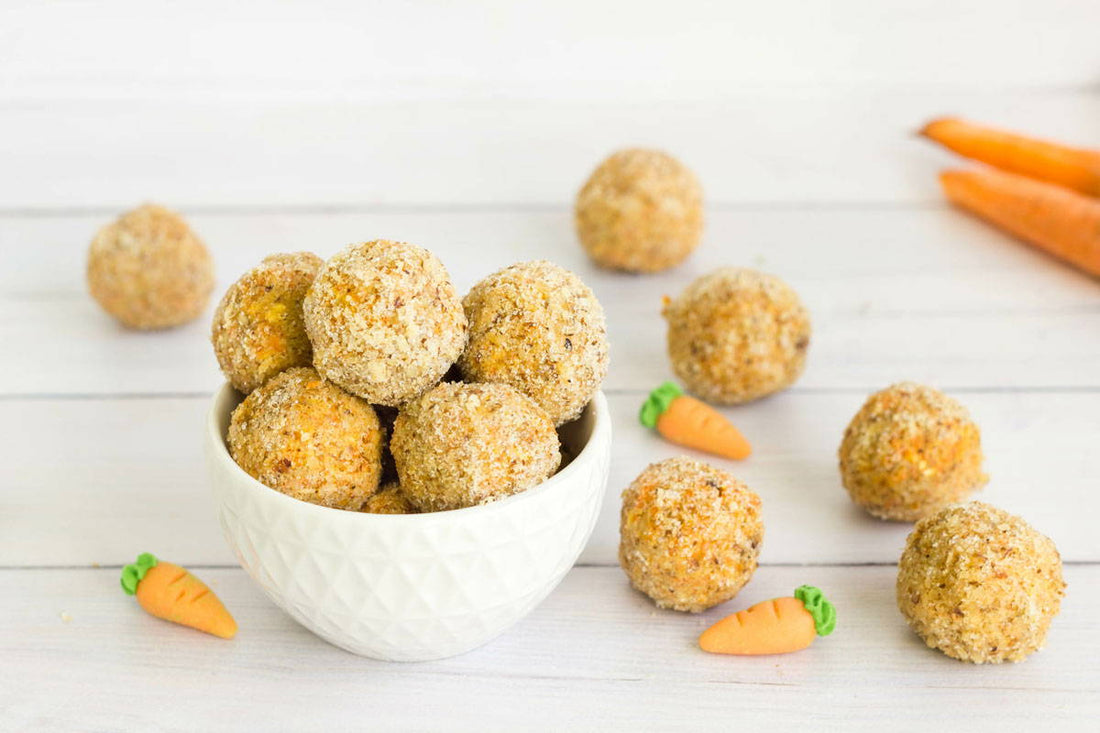 A white bowl filled with round, crumb-coated snacks sits on a light wooden surface. More snacks and small decorative carrots are scattered around the bowl. Two real carrots are partially visible in the background.