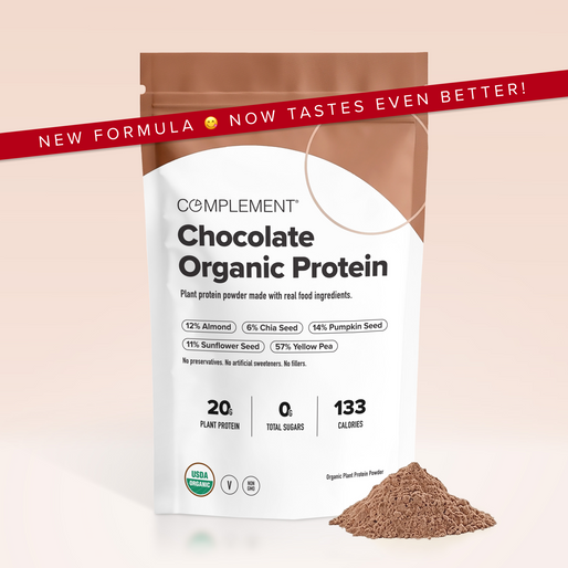 A bag of Complement Chocolate Organic Protein powder stands upright with some powder beside it. The bag highlights plant-based ingredients and protein content, with a red banner reading NEW FORMULA 😊 NOW TASTES EVEN BETTER!.