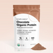 A pouch of Complement Chocolate Organic Protein powder stands upright, with some powder spilled beside it. The packaging highlights its plant-based ingredients and nutritional facts, including 15g protein per serving.