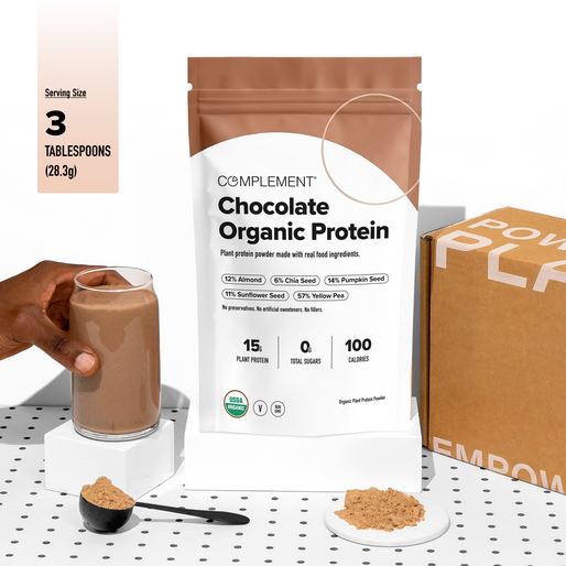 A hand holds a glass of chocolate protein shake next to a Complement Chocolate Organic Protein pouch, a spoonful of powder, and a box on a white, dotted surface. Nutritional info and serving size are shown.