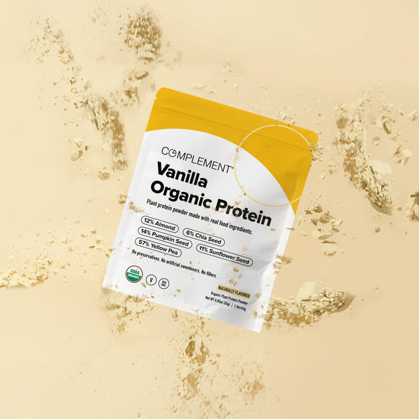 A packet of Complement Vanilla Organic Protein powder lies on a surface, surrounded by scattered protein powder. The packaging highlights plant-based ingredients and certifications.