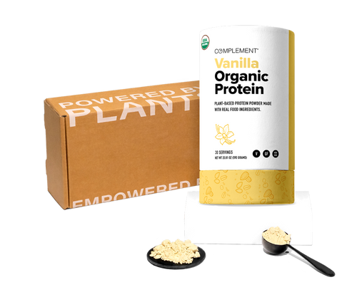 A container of Vanilla Organic Protein powder by Complement sits next to a cardboard box labeled Powered by Plants. There are two servings of protein powder, one on a black dish and another in a scoop.