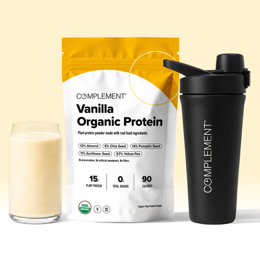 A glass of vanilla protein shake sits next to a Complement Vanilla Organic Protein powder bag and a black shaker bottle on a light surface with a yellow background accent.