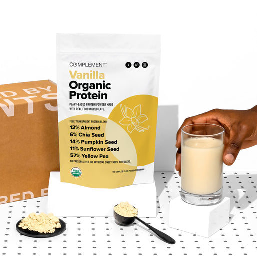 A bag of Complement Vanilla Organic Protein powder is displayed next to a glass of protein shake, a scoop of powder, and a black spoon. A cardboard box is in the background on a white surface with black dots.