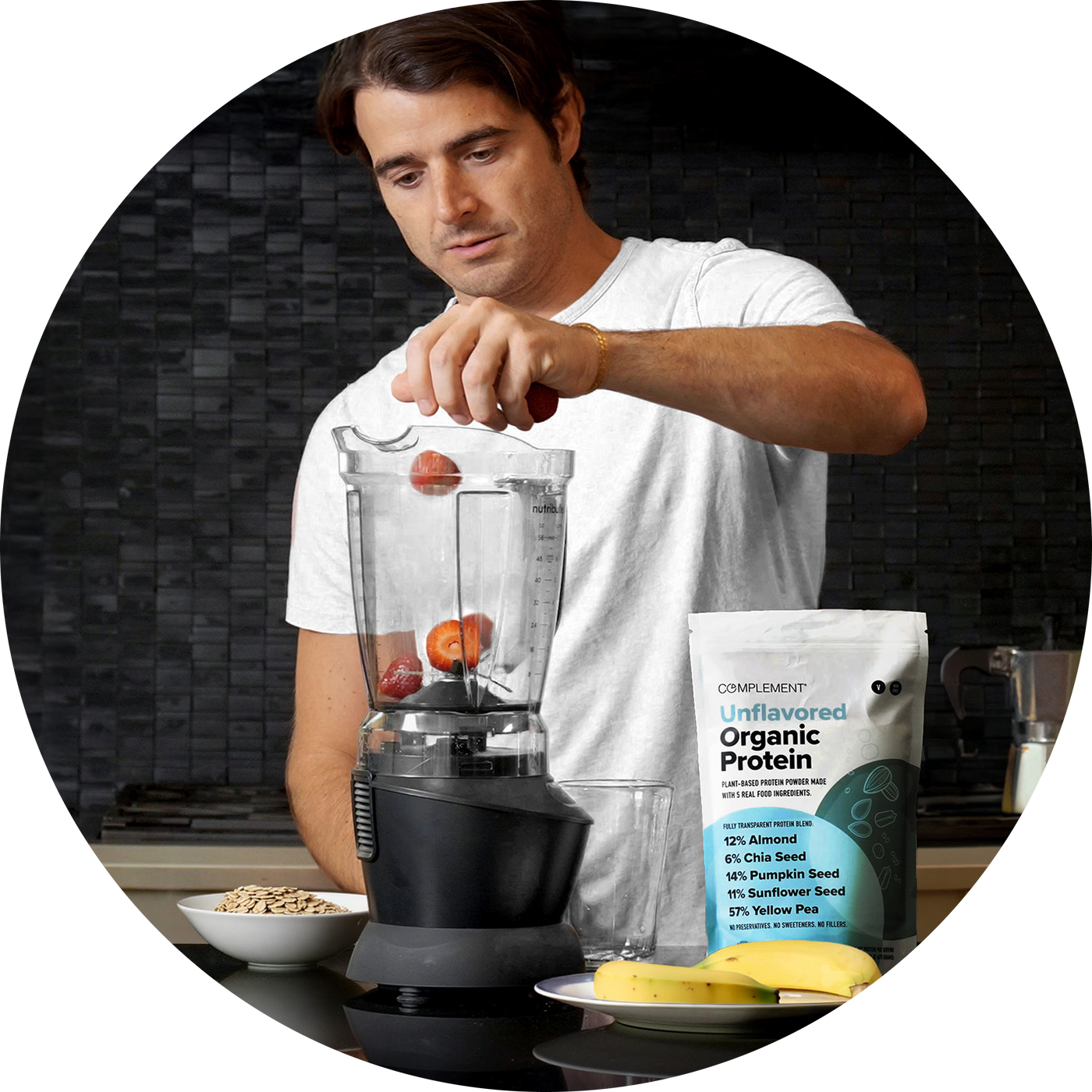 A man in a white shirt adds strawberries to a blender in a kitchen. Beside him are a packet of organic protein powder, a bowl of seeds, and some bananas on the counter.