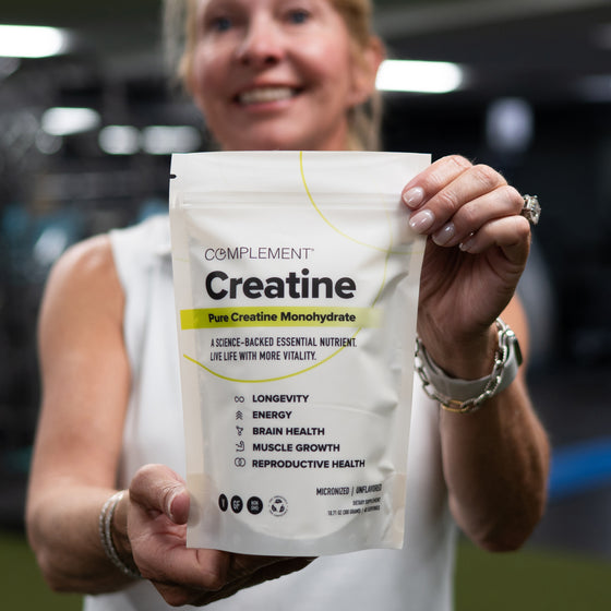 A smiling person in a white sleeveless top holds a package of Complement Creatine Monohydrate, emphasizing benefits like longevity, energy, brain health, muscle growth, and reproductive health.