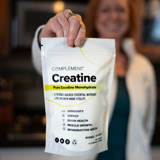 A person holds a white bag of Complement Creatine Monohydrate supplement toward the camera. The packaging highlights benefits like longevity, energy, brain health, muscle growth, and reproductive health.