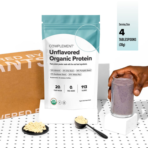 A hand holds a glass of purple protein smoothie next to a packet of Complement Unflavored Organic Protein and two measuring spoons with protein powder on a polka dot surface.