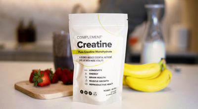 A package of Complement Creatine on a kitchen counter. The bag highlights benefits like longevity and energy. Nearby are strawberries and bananas on a cutting board, with blurred background elements including a milk bottle.